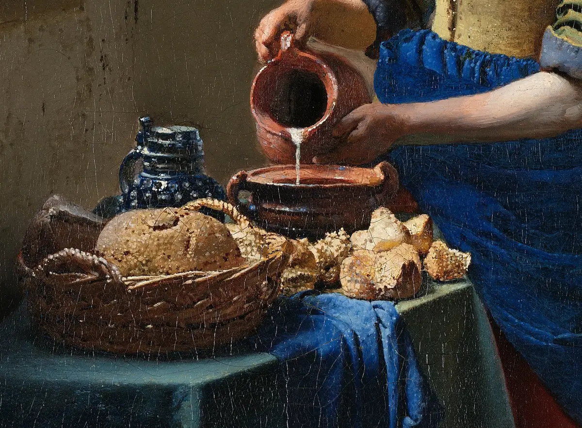 Detail of the table: a wicker basket of bread rolls, a blue and white jug, and milk pouring from a red earthenware jug into a bowl.
